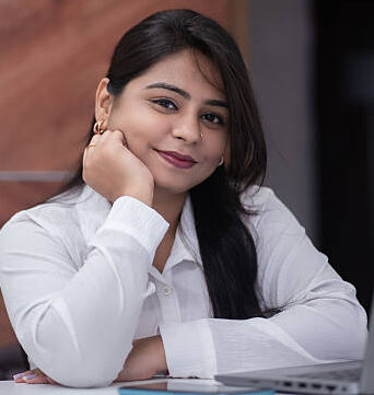 Portrait of a cheerful young Indian woman in formal attire smiling at the camera with her chin resting on her hands. Seated at a work desk with a laptop, phone, and coffee mug in view, this image portrays a positive and confident professional in a modern indoor office setting.